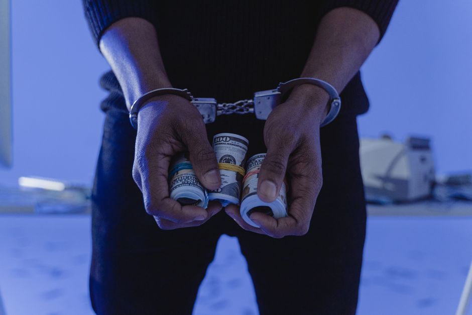 A person wearing dark clothing and handcuffs is holding several rolled-up banknotes, including denominations of hundred-dollar bills, with their fingers wrapped around the currency. The background fea