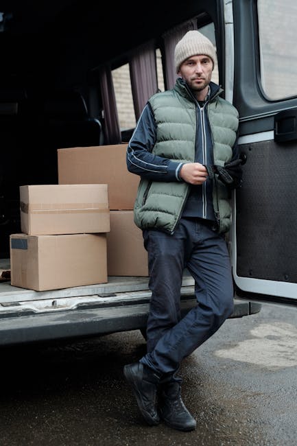 A man dressed in casual clothing, including a black and blue jacket, black trousers, and white footwear, stands next to the open rear door of a white moving van parked on a driveway in front of a resi